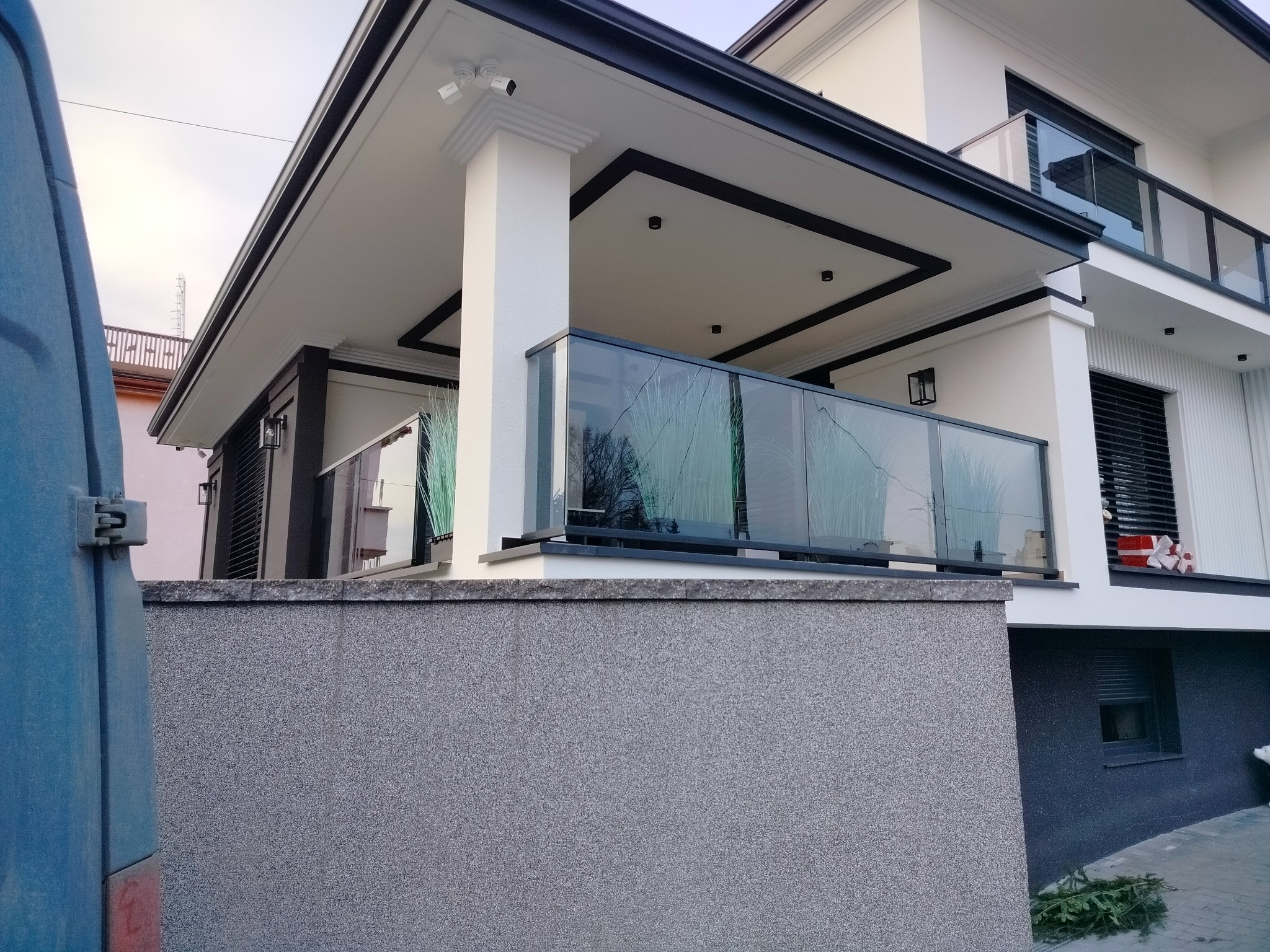 Nowoczesny dom z balkonem zabezpieczonym szkłem hartowanym, widok z dołu na balustradę z delikatnym wzorem drzewa odbijającym się na szkle, z boku fragment niebieskiego samochodu dostawczego.