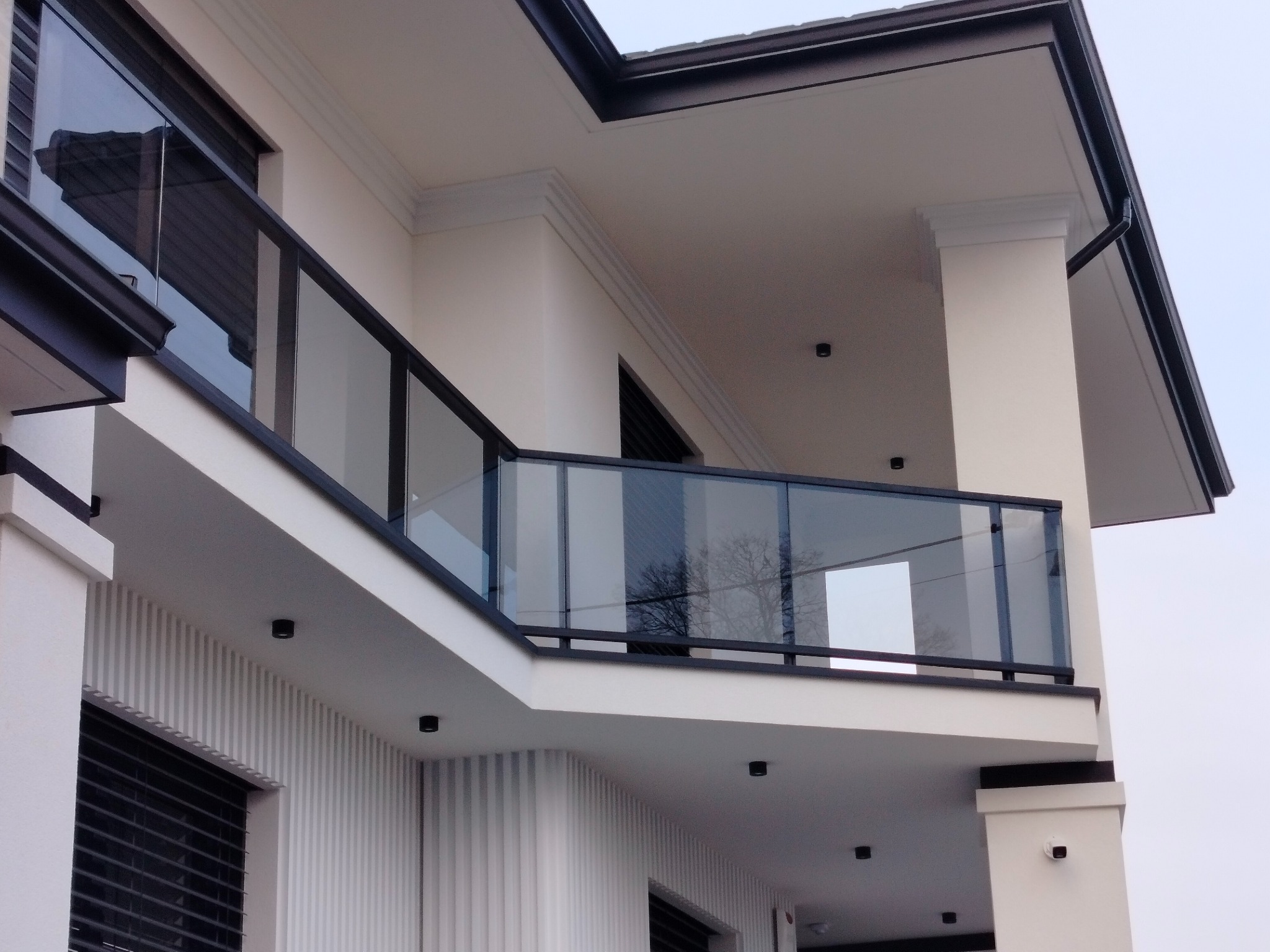 Nowoczesna szklana balustrada balkonowa z czarnymi profilami, widok z dołu na elewację budynku z oświetleniem punktowym.