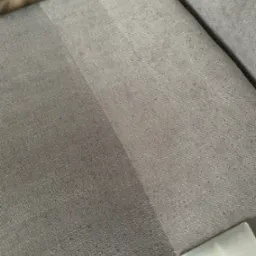 Fragment szarej kanapy z widocznym efektem czyszczenia, gdzie połowa powierzchni jest wyraźnie jaśniejsza po praniu, a w rogu kadru widoczna dysza odkurzacza piorącego.