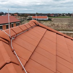 KWR Perun - Czerwona dachówka ceramiczna z zamontowanym systemem ochrony odgromowej, widok z góry na tle zabudowań i pola. Metalowe elementy mocujące.