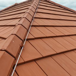 KWR Perun - Czerwona dachówka ceramiczna z zamontowanym przewodem odprowadzającym, zabezpieczającym budynek przed wyładowaniami atmosferycznymi. Widok z góry, pochmurne niebo.