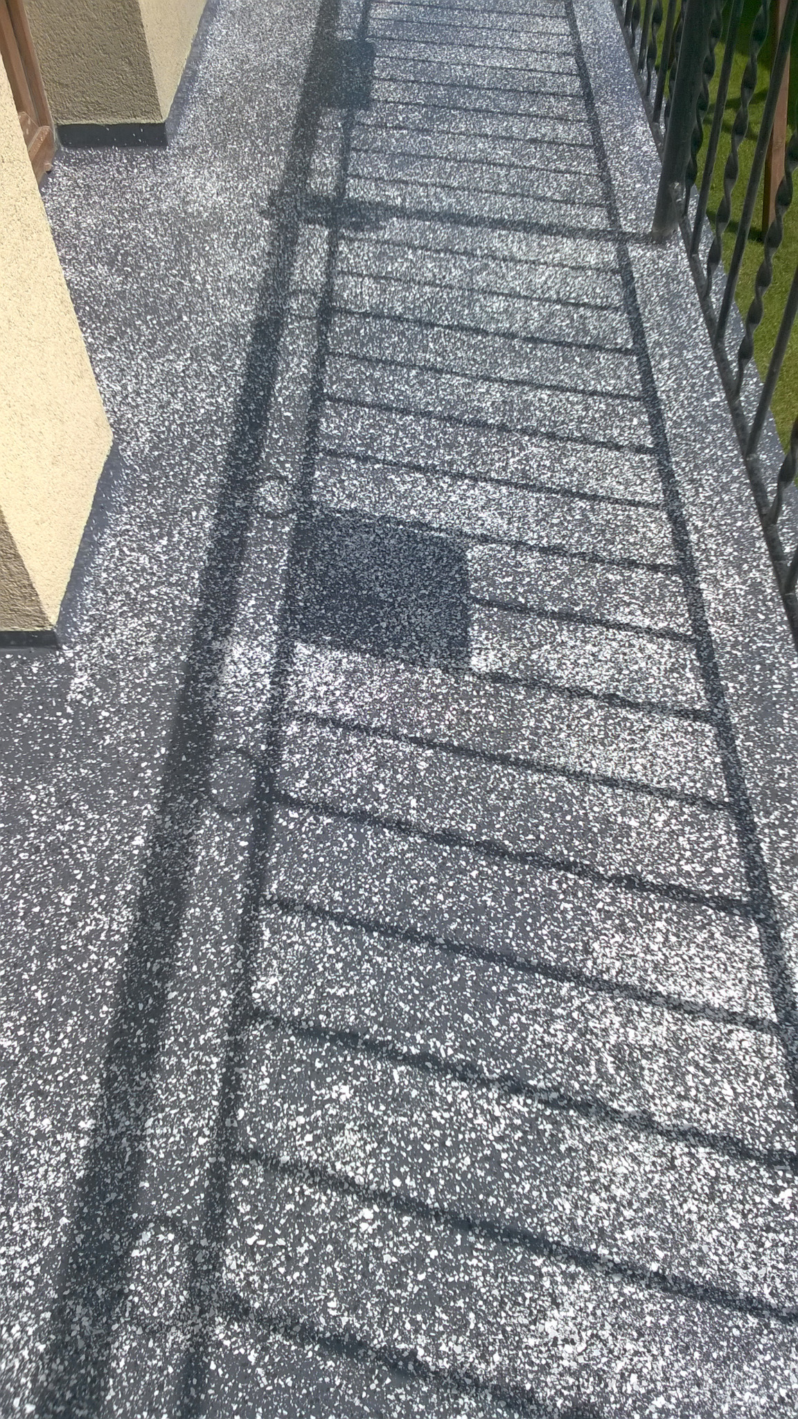 Szara, drobnoziarnista powierzchnia balkonu z balustradą w tle, oświetlona słońcem, rzucająca cień kratownicy.