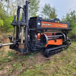 Pomarańczowo-czarna wiertnica kierunkowa Ditch Witch JT30 na gąsienicach, przygotowana do pracy na zewnątrz; widoczne przewody rurowe i błotniste podłoże.