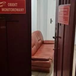 Wnętrze kancelarii komorniczej: widok na poczekalnię z brązową sk&oacute;rzaną sofą, przez uchylone drewniane drzwi z tabliczką informacyjną o monitoringu i danymi komornika sądowego.