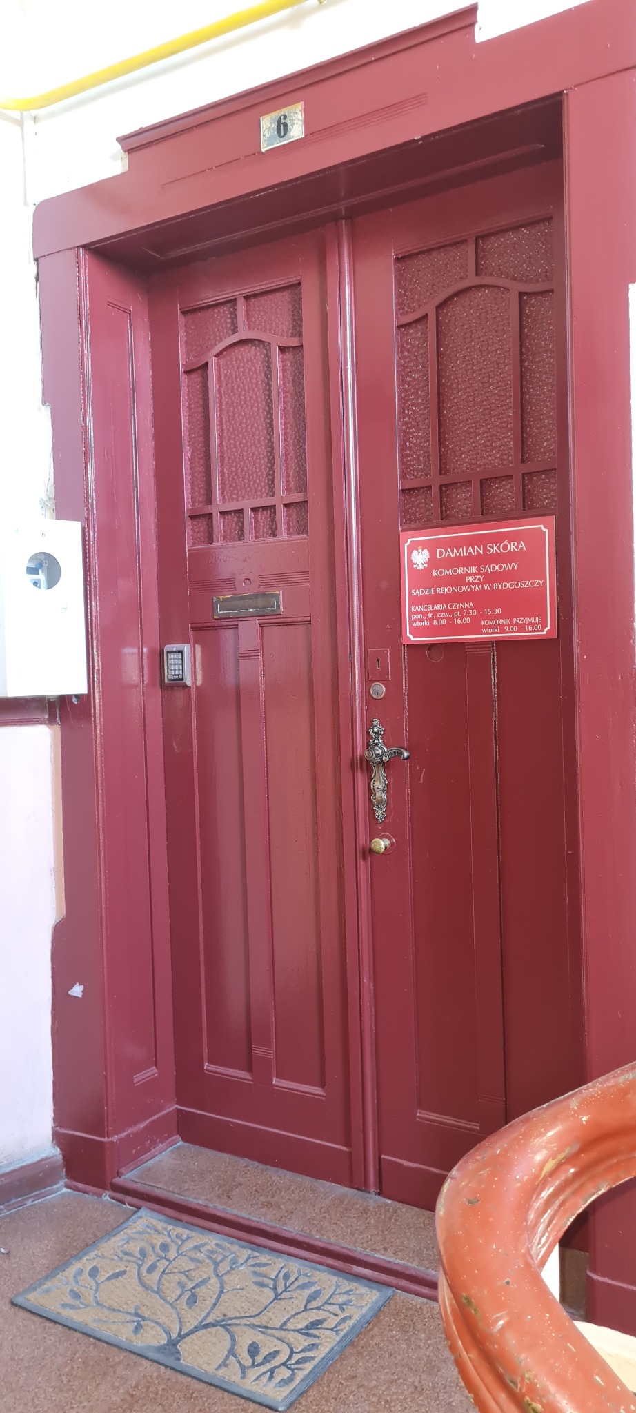 Wejście do kancelarii komornika sądowego Damiana Skóry w Bydgoszczy, czerwone dwuskrzydłowe drzwi z szyldem informacyjnym o godzinach urzędowania.