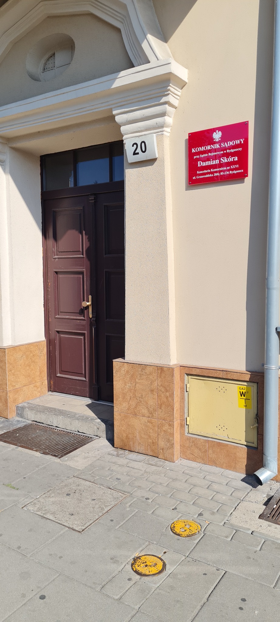 Wejście do kancelarii komornika sądowego Damiana Skóry przy ulicy Grunwaldzkiej 20/6 w Bydgoszczy, z widoczną tabliczką informacyjną i numerem 20 nad brązowymi drzwiami