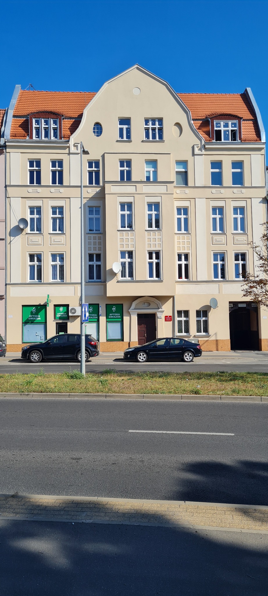 Elewacja budynku z apteką na parterze, widoczne okna z logotypami i zaparkowane samochody wzdłuż ulicy.
