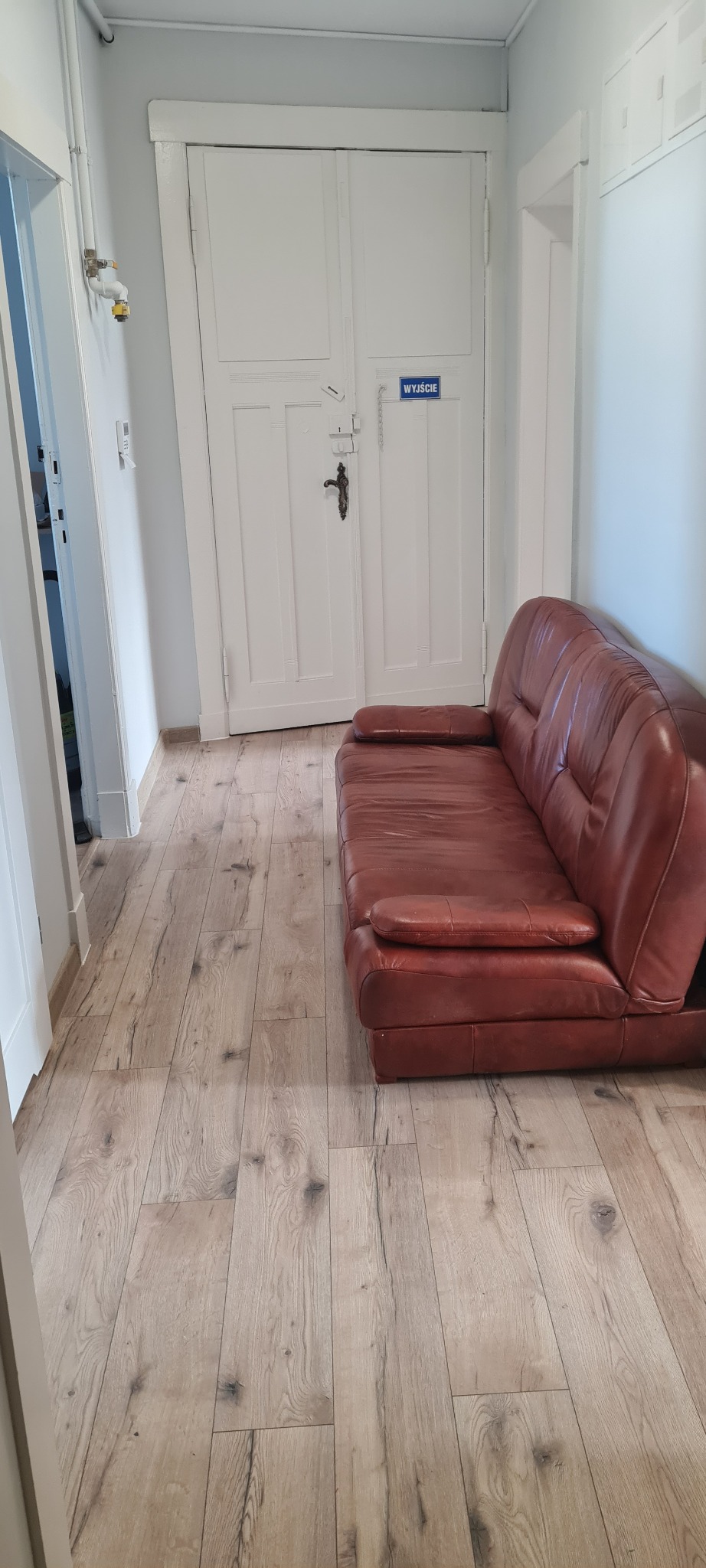 Czerwona skórzana sofa ustawiona w korytarzu z drewnianą podłogą, na tle białych drzwi z napisem 'Wyjście'.
