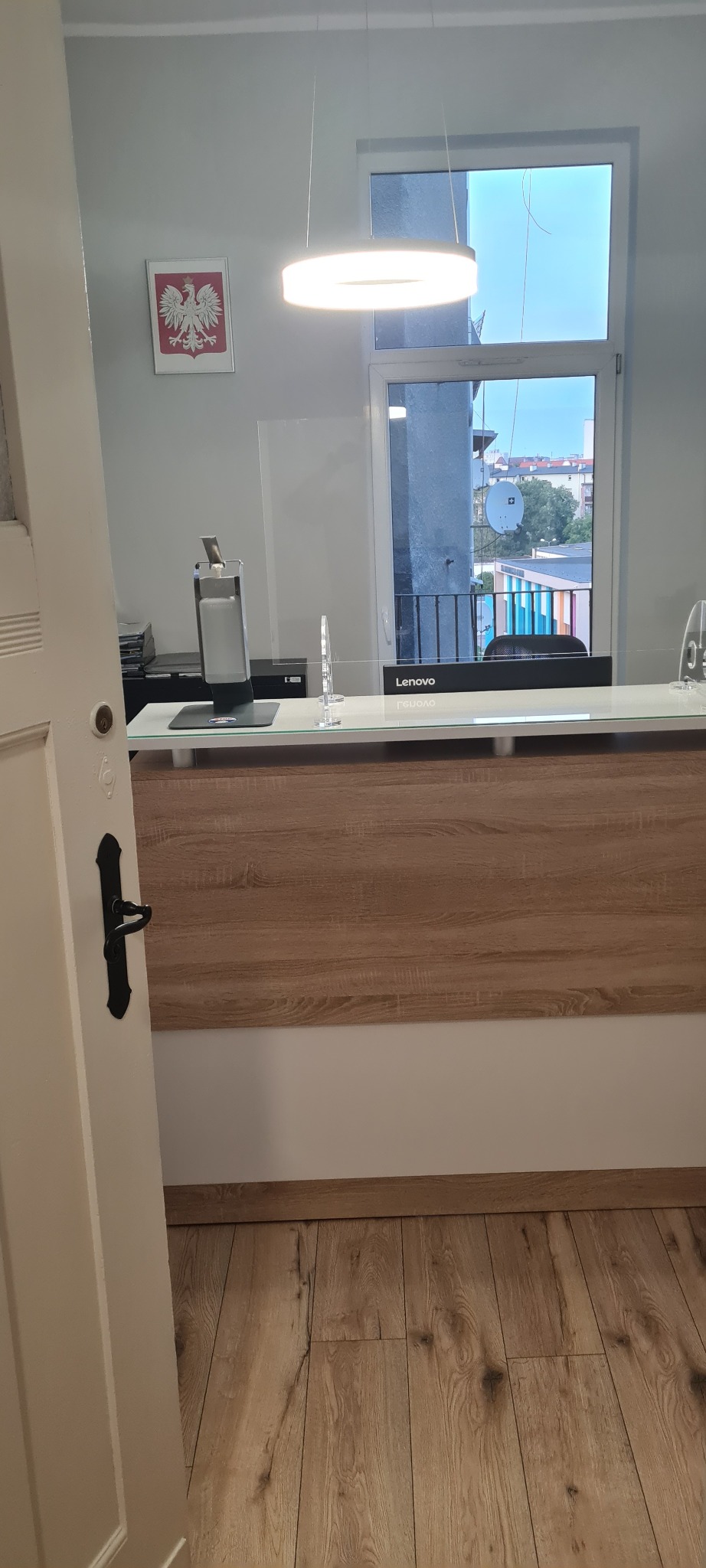 Biuro z recepcją zabezpieczoną szybą ochronną, widoczny monitor Lenovo, na ścianie godło Polski, widok z okna na miasto.