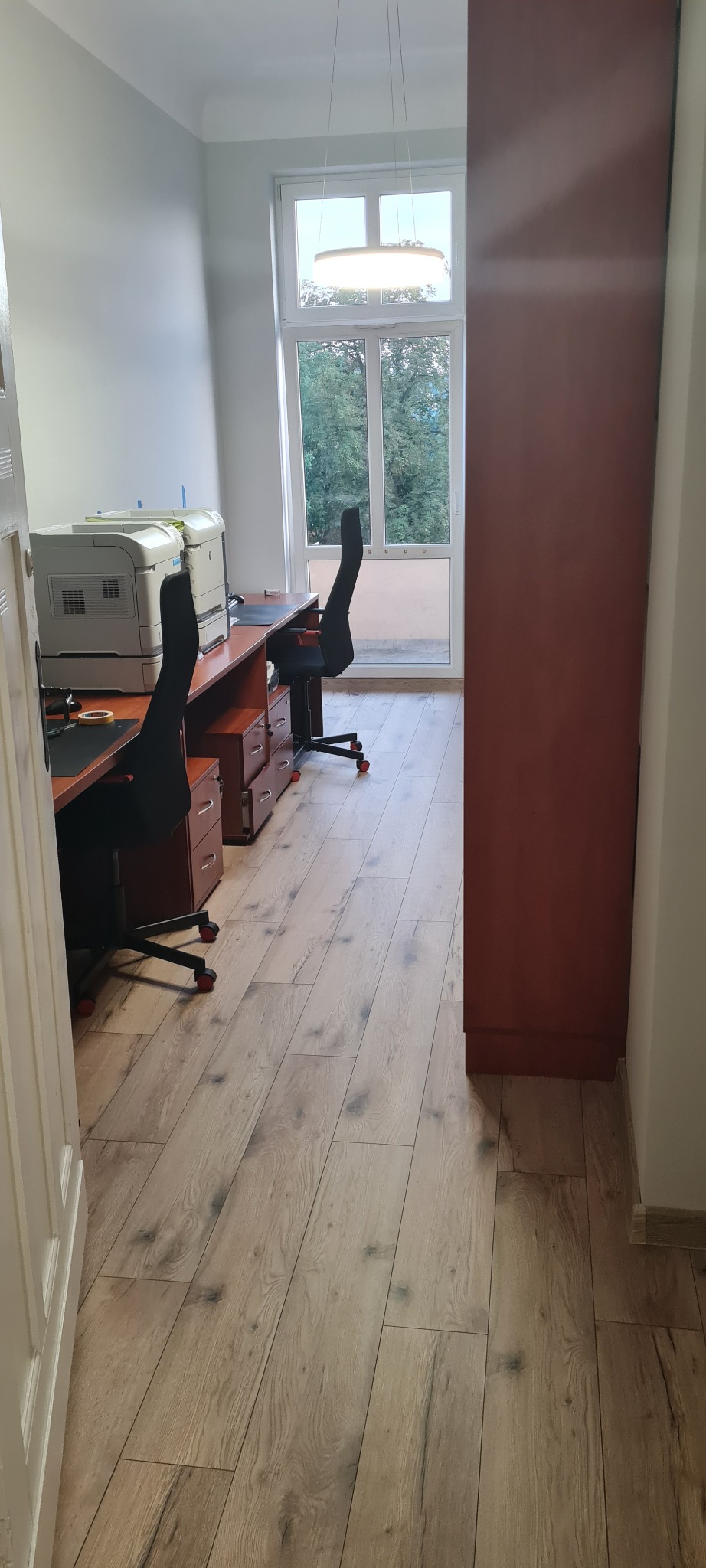 Biuro z dwoma stanowiskami pracy, biurkami z szufladami i krzesłami obrotowymi, drukarki na biurkach, widok na zieleń za oknem.