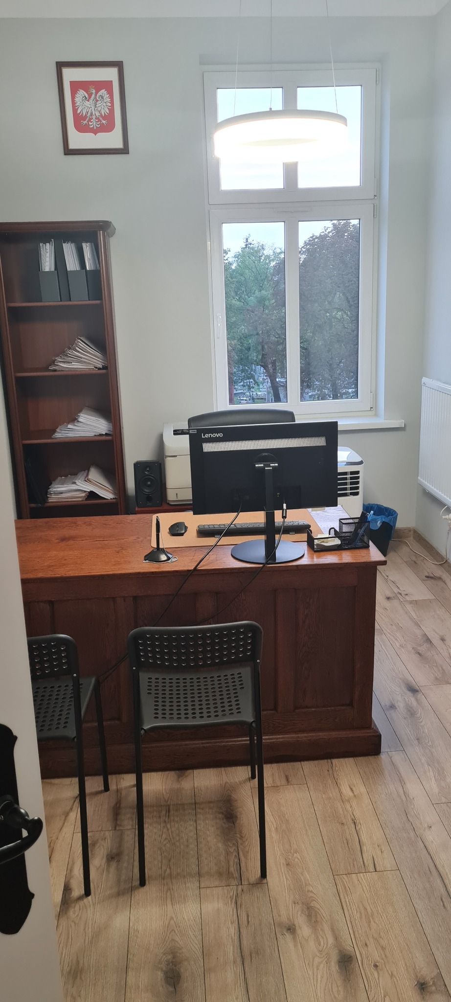 Biuro z drewnianym biurkiem, komputerem Lenovo, regałem na dokumenty i godłem Polski na ścianie.