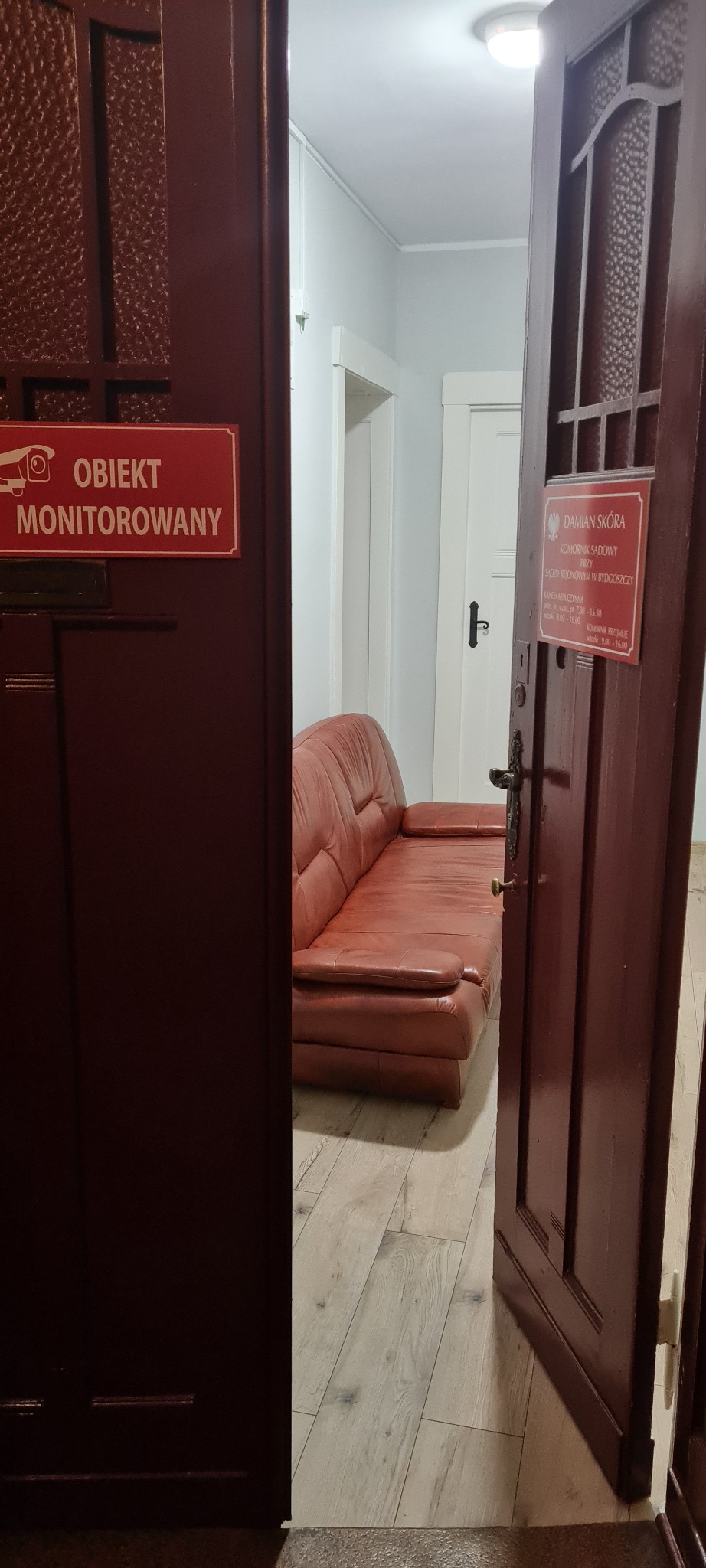 Wnętrze kancelarii komorniczej: widok na poczekalnię z brązową skórzaną sofą, przez uchylone drewniane drzwi z tabliczką informacyjną o monitoringu i danymi komornika sądowego.