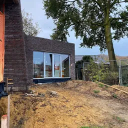 Budynek w trakcie budowy z cegły klinkierowej, z zamontowanymi oknami z białymi ramami, widok z zewnątrz na plac budowy z wykopem i materiałami budowlanymi.