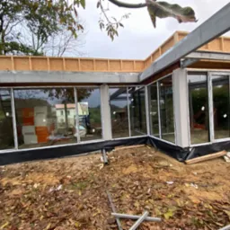 Budowa przeszklonego aneksu domu w trakcie realizacji, widoczne okna z białymi ramami, konstrukcja dachu i elementy wykończeniowe, teren budowy z liśćmi i narzędziami.