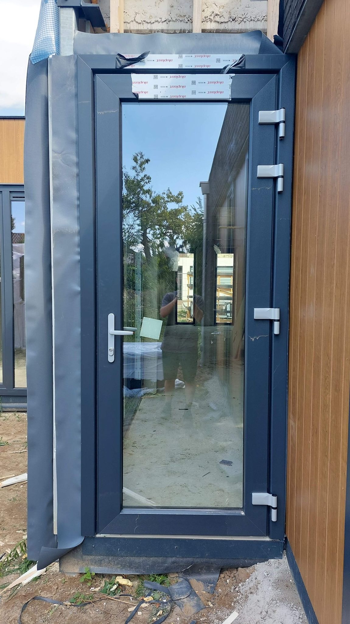 Zamontowane szare drzwi balkonowe z widocznymi zawiasami, uszczelkami i folią ochronną na ramie, odbijające otoczenie w szybie.