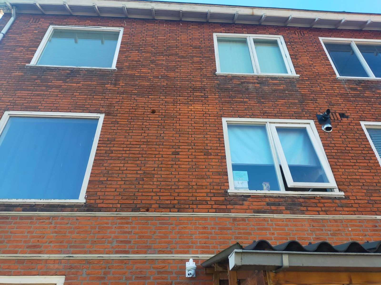 Widok z dołu na fasadę budynku z czerwonej cegły we Wrocławiu, z kilkoma białymi oknami, jedno z nich uchylone, widoczna kamera monitoringu na ścianie.