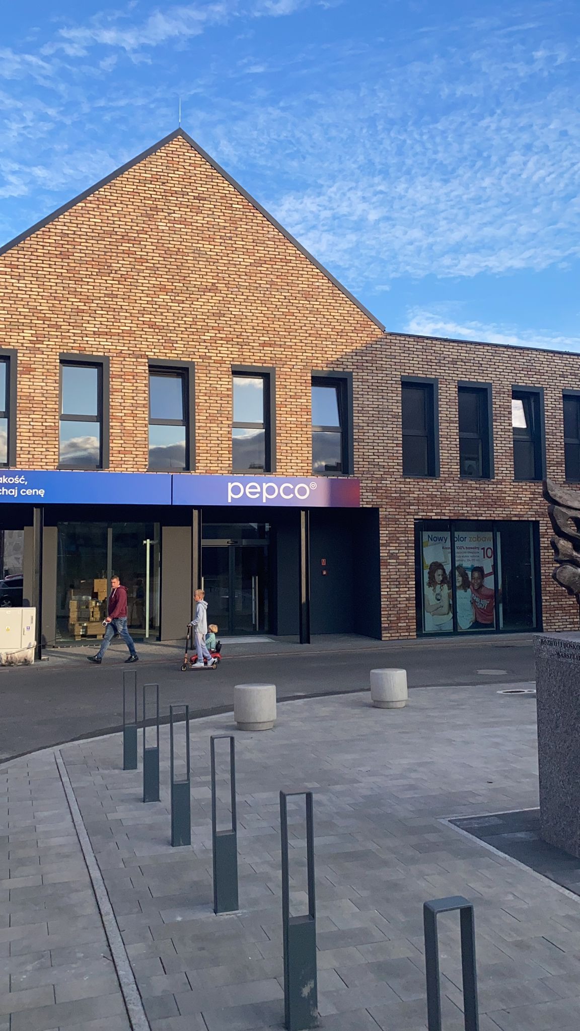 Fasada budynku sklepu Pepco z cegły, z czarnymi oknami i drzwiami, na tle błękitnego nieba z delikatnymi chmurami. Przed budynkiem przechodzą ludzie i stoi pomnik.