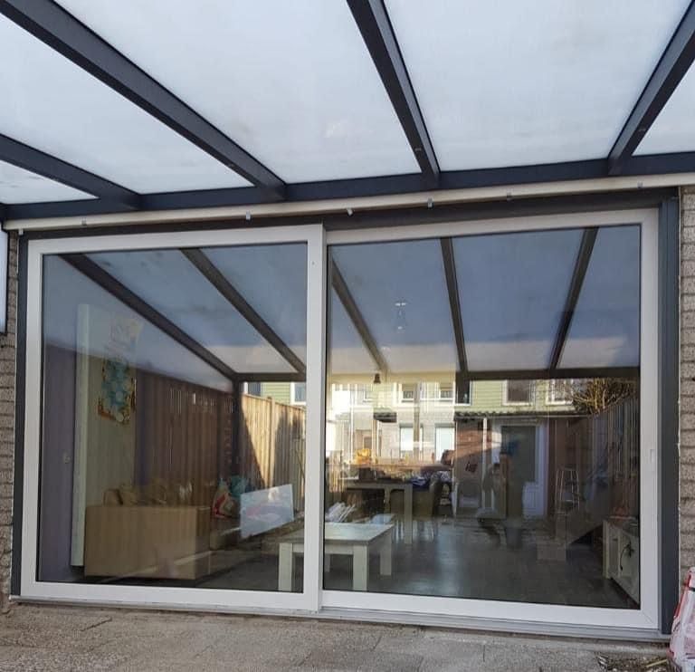 Przesuwne, białe drzwi tarasowe z dużym przeszkleniem, widok na zadaszony taras z meblami ogrodowymi i zewnętrzny ogród.