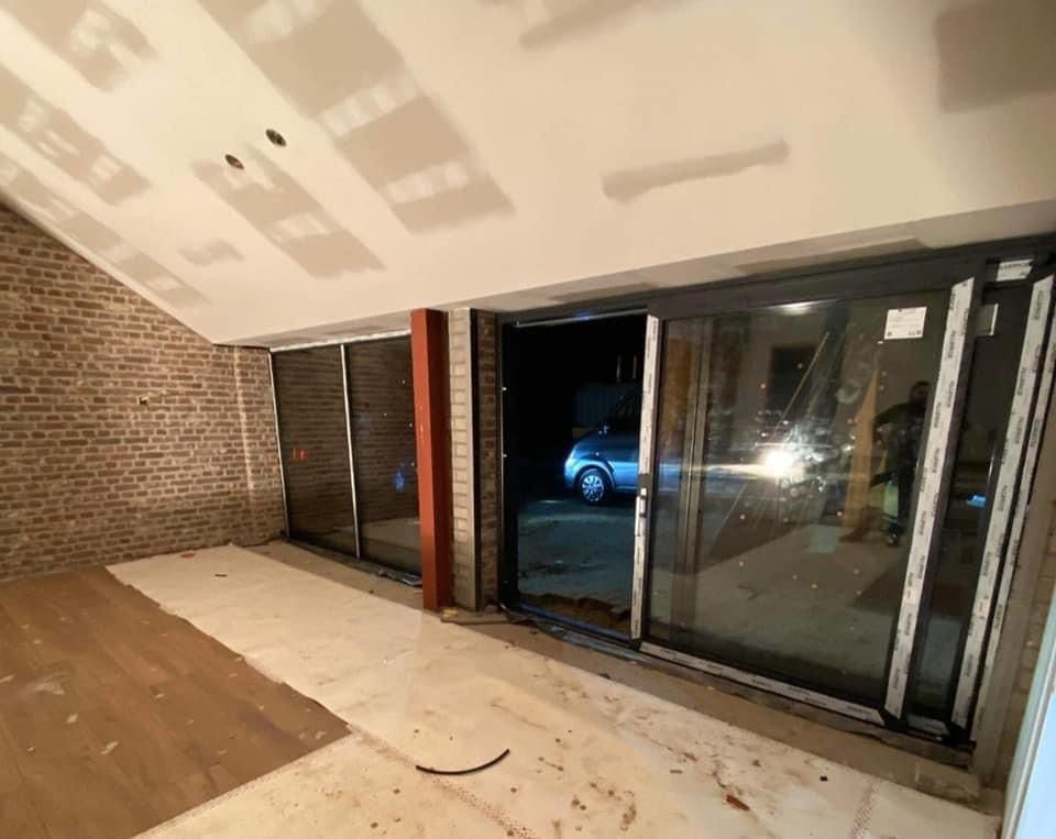 Surowy stan wnętrza z zamontowanymi przesuwnymi oknami balkonowymi z ciemnymi ramami, widoczne ślady prac wykończeniowych na podłodze i ścianach, przez okno widoczny samochód w nocy.