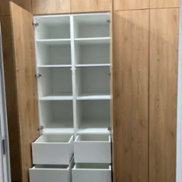 Wnętrze szafy wnękowej z otwartymi drzwiami, drewnopodobne fronty, białe półki i szuflady, widoczne zawiasy.