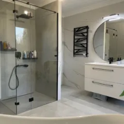 Nowoczesna łazienka z kabiną prysznicową typu walk-in z czarnymi okuciami, okrągłym lustrem z podświetleniem, białą szafką pod umywalkę i grzejnikiem drabinkowym w geometryczny wzór.