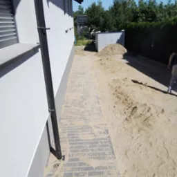 Świeżo ułożona, szara kostka brukowa przy białej ścianie domu z czarną rynną, widoczny piasek i ziemia przygotowana pod dalsze prace.