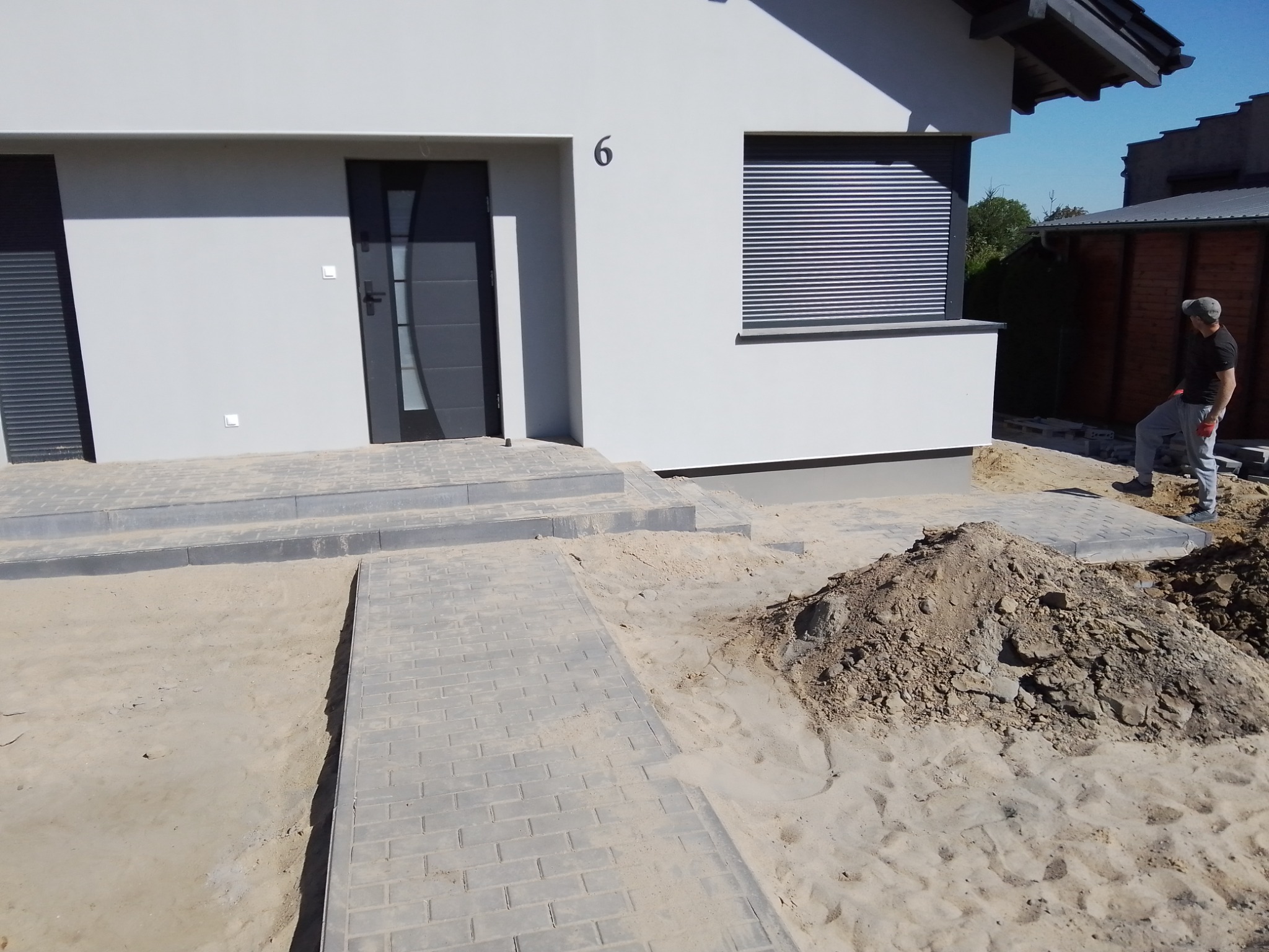 Nowoczesny dom z jasną elewacją i numerem 6, z częściowo ułożoną ścieżką z szarej kostki brukowej prowadzącą do wejścia, obok hałda ziemi i pracownik w tle.