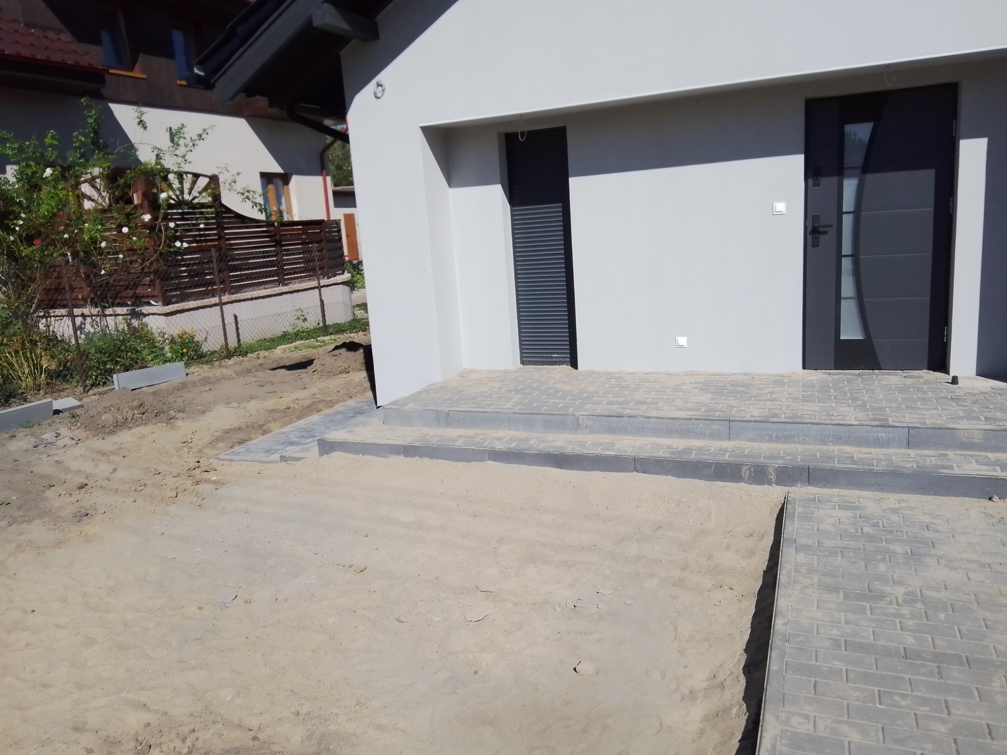 Niedokończone podejście do domu z kostki brukowej, widoczne schody i przygotowane podłoże z piasku.