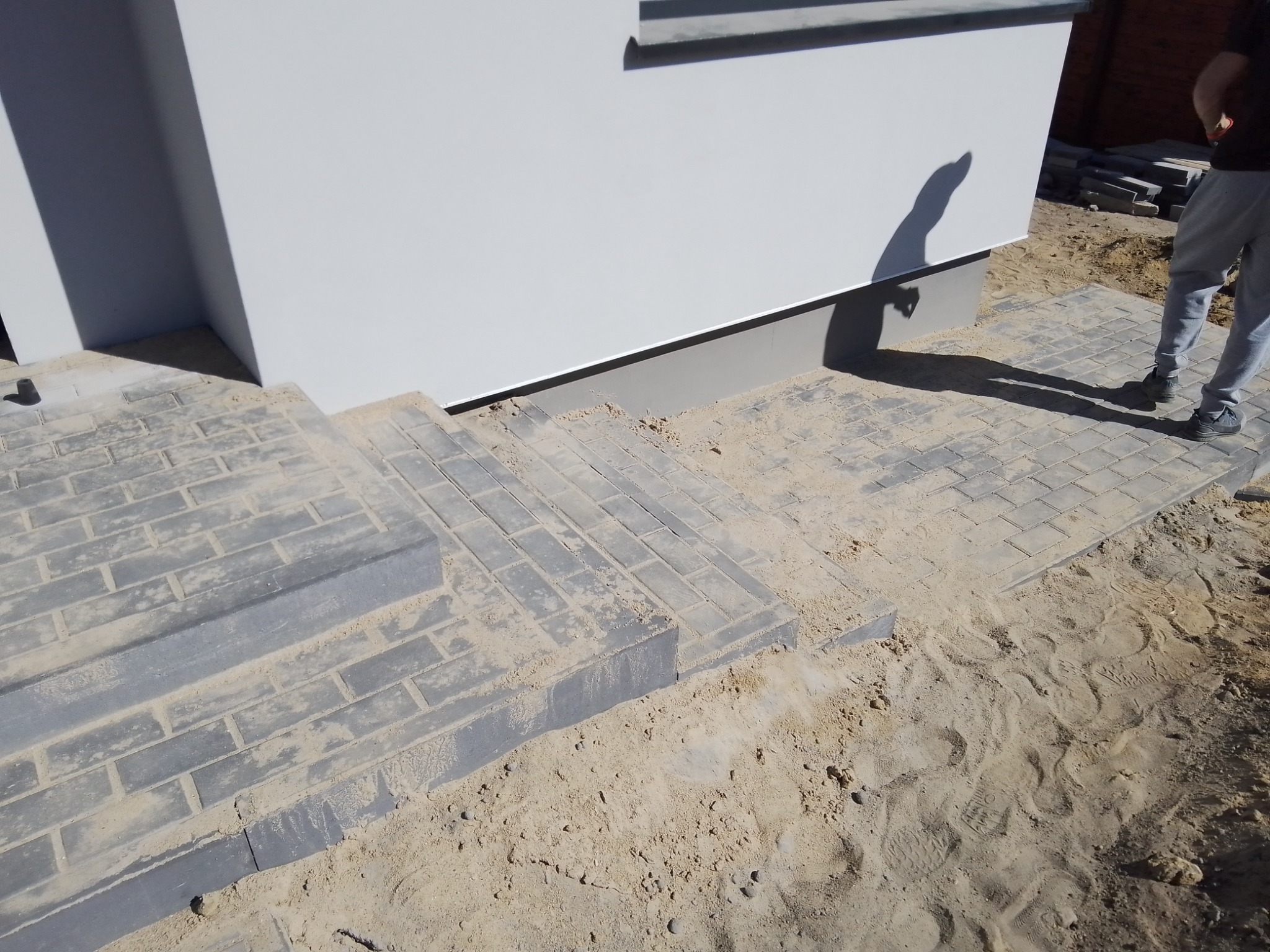 Schody i chodnik z szarej kostki brukowej w trakcie budowy, widoczny piasek zasypujący szczeliny, cień osoby na ścianie budynku.