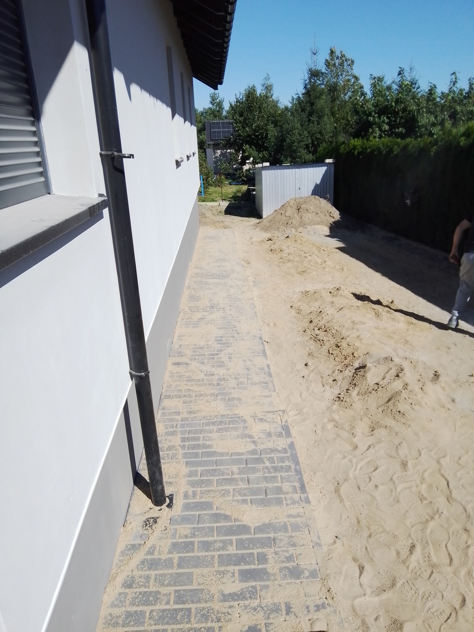 Świeżo ułożona, szara kostka brukowa przy białej ścianie domu z czarną rynną, widoczny piasek i ziemia przygotowana pod dalsze prace.