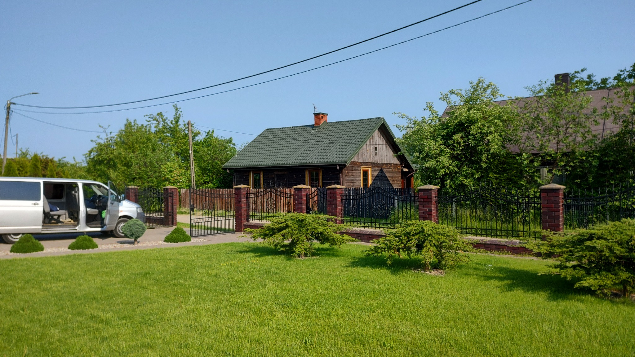 Zadbany trawnik przed drewnianym domem z zielonym dachem, ozdobne krzewy iglaste, metalowe ogrodzenie z ceglanymi słupkami, srebrny van z otwartymi drzwiami po lewej stronie.