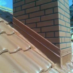 Wykończony komin z cegły klinkierowej wystający ponad połać dachu pokrytą ceramiczną dachówką, obróbka blacharska komina z uszczelnieniem dekarskim.