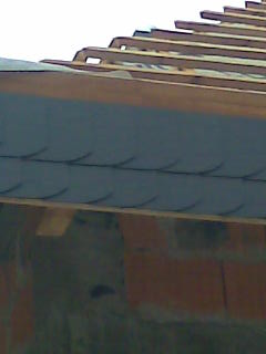 Fragment dachu w trakcie budowy, widoczne szare płytki układane na drewnianej konstrukcji, mur z cegły w tle.