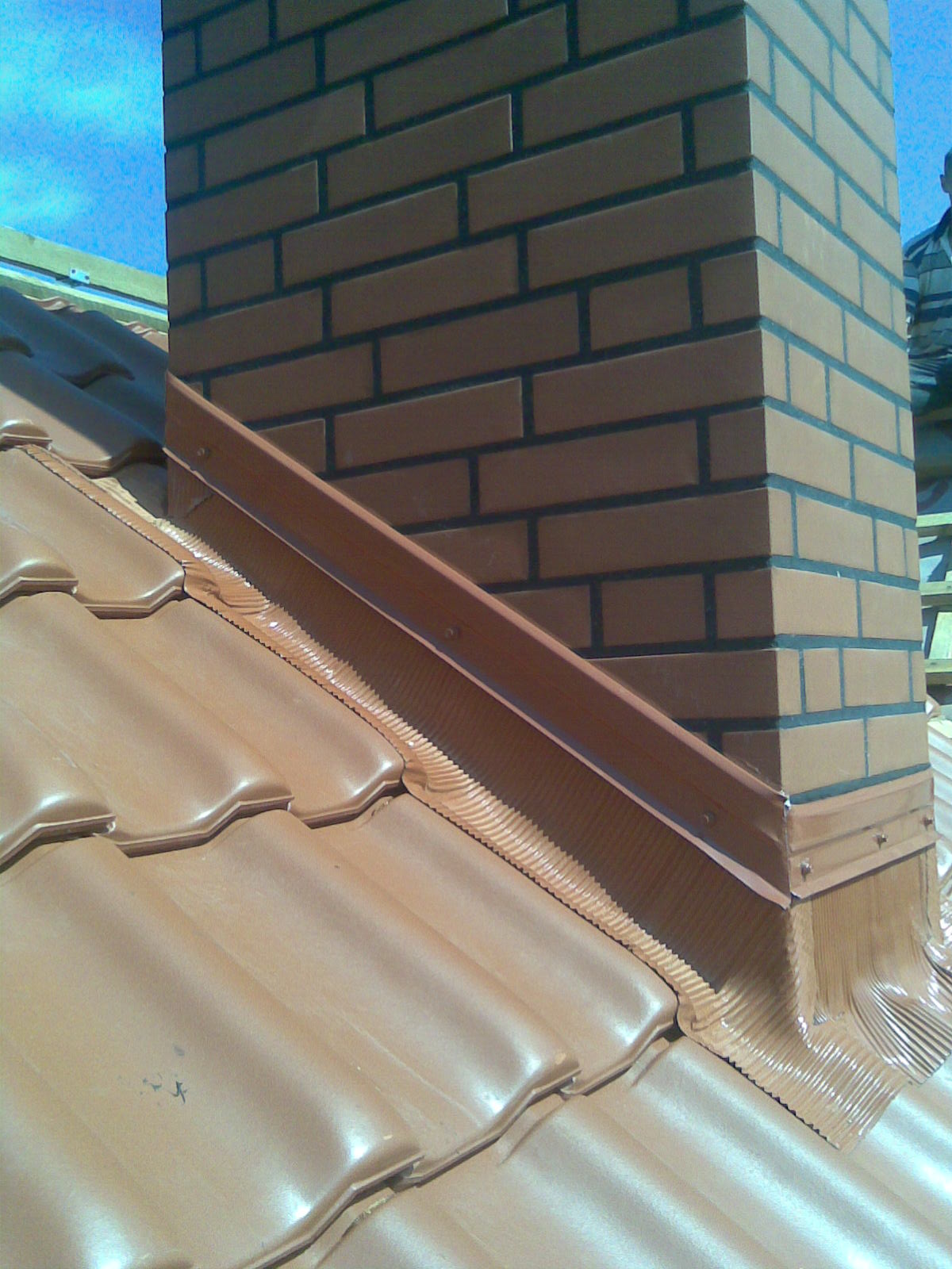 Wykończony komin z cegły klinkierowej wystający ponad połać dachu pokrytą ceramiczną dachówką, obróbka blacharska komina z uszczelnieniem dekarskim.