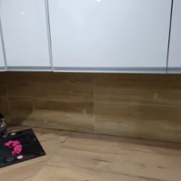 Nowoczesna kuchnia z białymi szafkami wiszącymi, drewnopodobnymi panelami ściennymi i podłogą, czarnym zlewem z chromowaną baterią i różową ścierką.