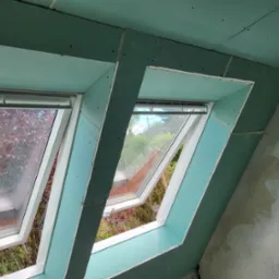 Dwa okna dachowe w trakcie montażu, widoczne płyty gipsowo-kartonowe i surowa ściana z odręcznymi notatkami.