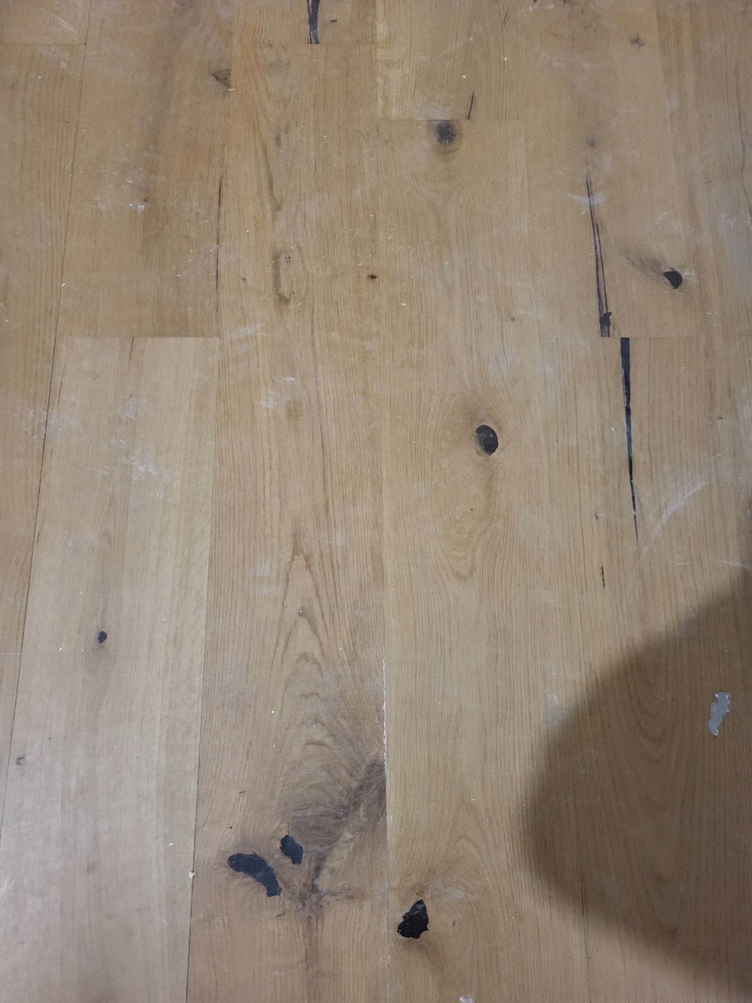 Położona drewniana podłoga z widocznymi sękami i drobnymi zabrudzeniami, uchwycona z góry, z cieniem w prawym dolnym rogu.
