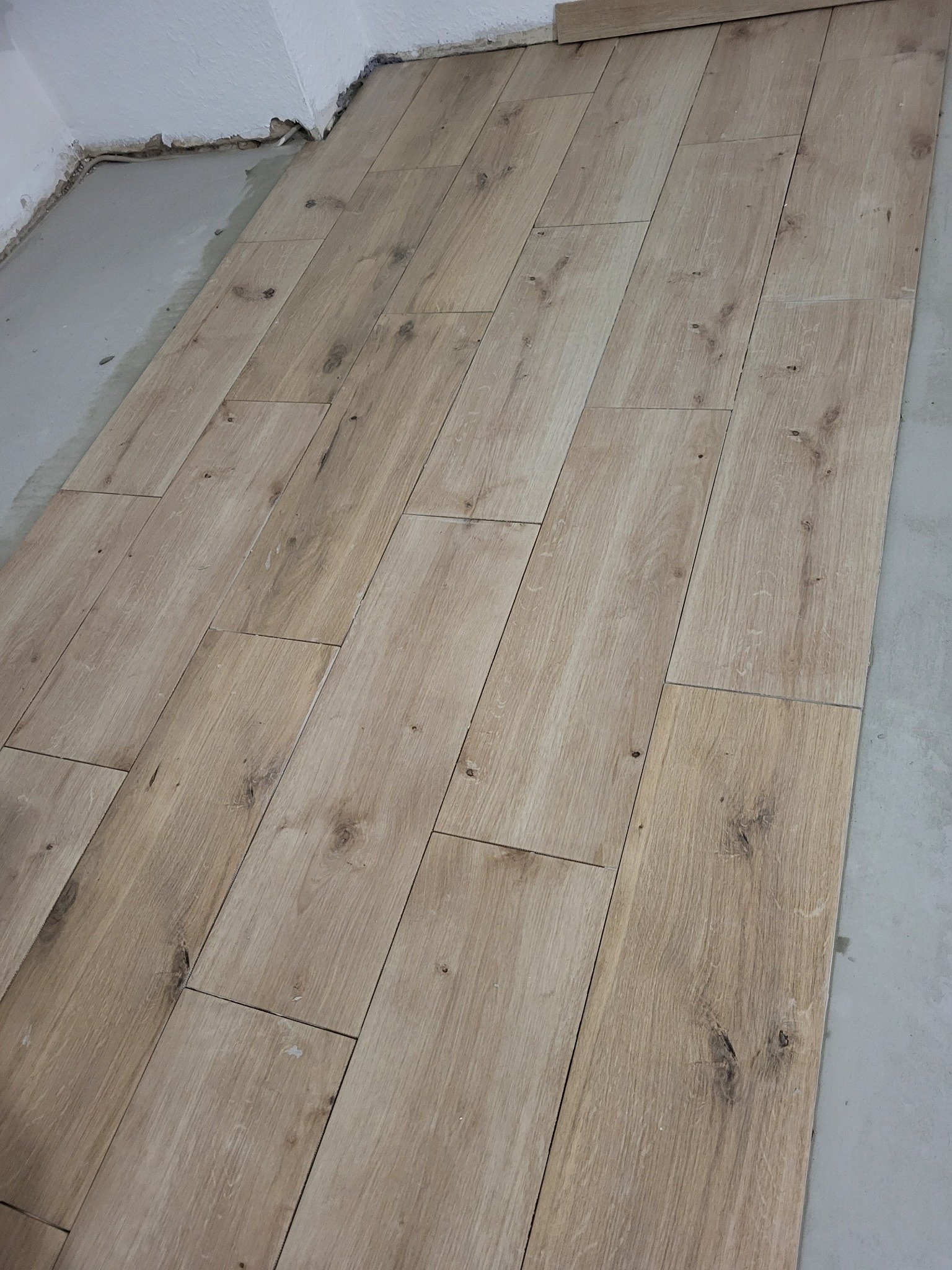 Układanie paneli podłogowych imitujących drewno, widoczne niedociągnięcia przy ścianie i surowy beton pod spodem.