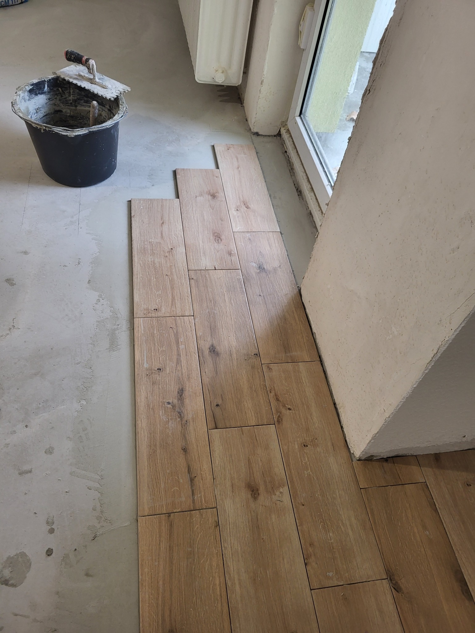 Układanie podłogi z płytek imitujących drewno, widoczny kubeł z zaprawą i kielnia, surowy stan podłoża, grzejnik i okno w tle.