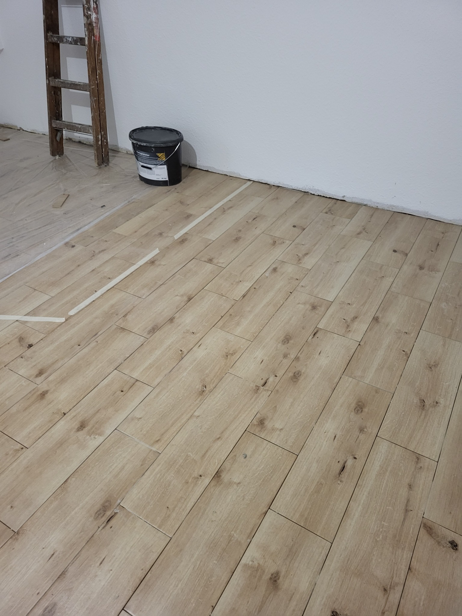 Podłoga wyłożona panelami imitującymi drewno, zabezpieczona folią malarską, drabina malarska i wiadro z farbą w rogu pomieszczenia, ślady taśmy malarskiej na panelach.