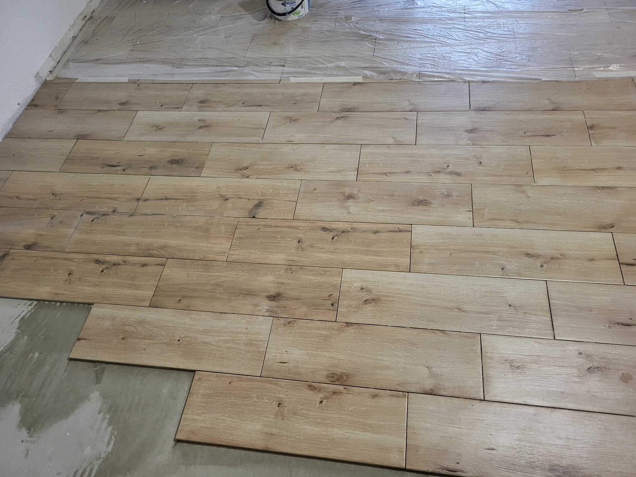 Układanie podłogi z płytek imitujących drewno w trakcie prac, widoczna folia ochronna na ścianie i wiadro z zaprawą.