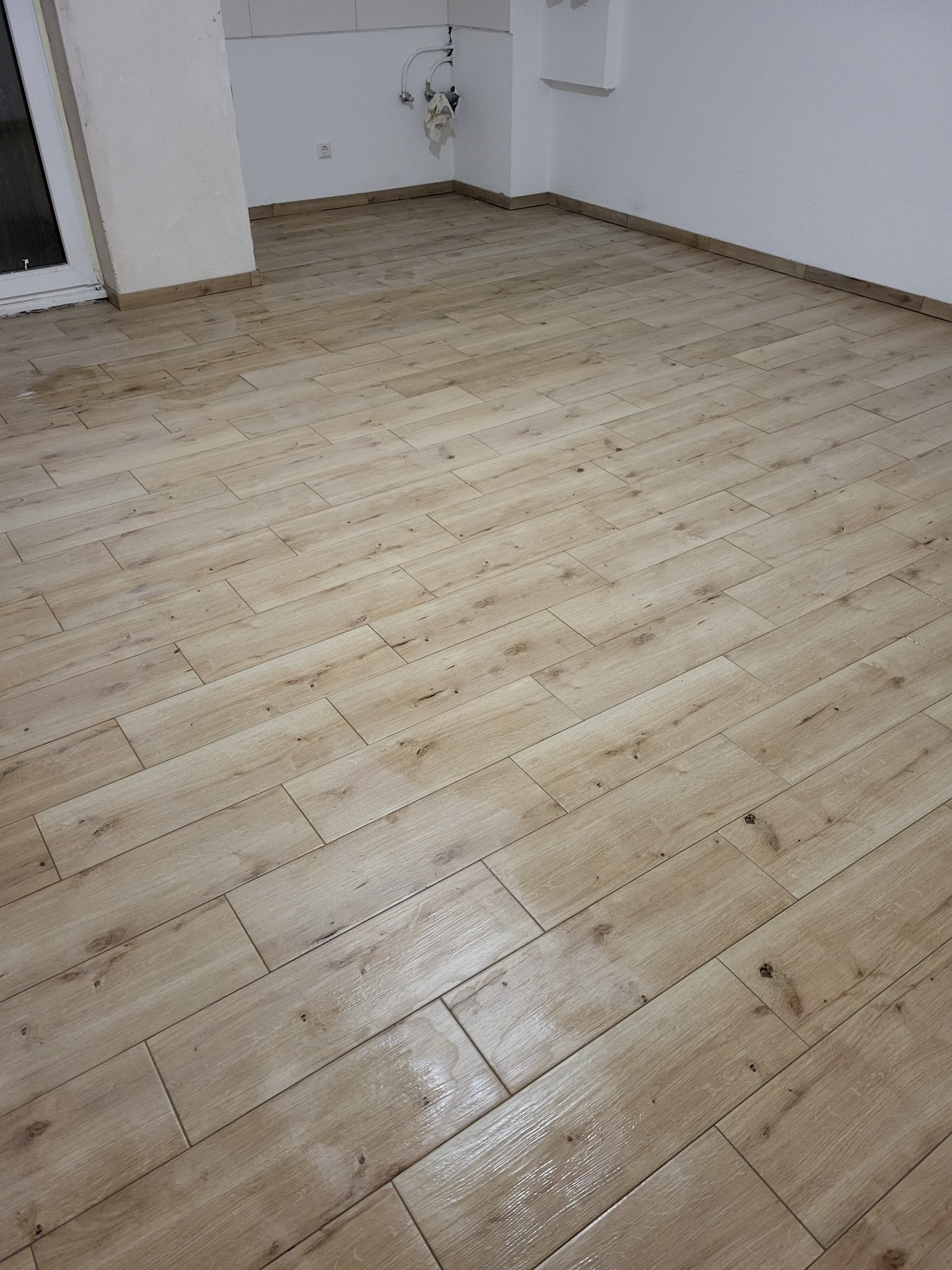 Ułożona podłoga z płytek imitujących drewno w pustym, jasnym pomieszczeniu z białymi ścianami i listwami przypodłogowymi.