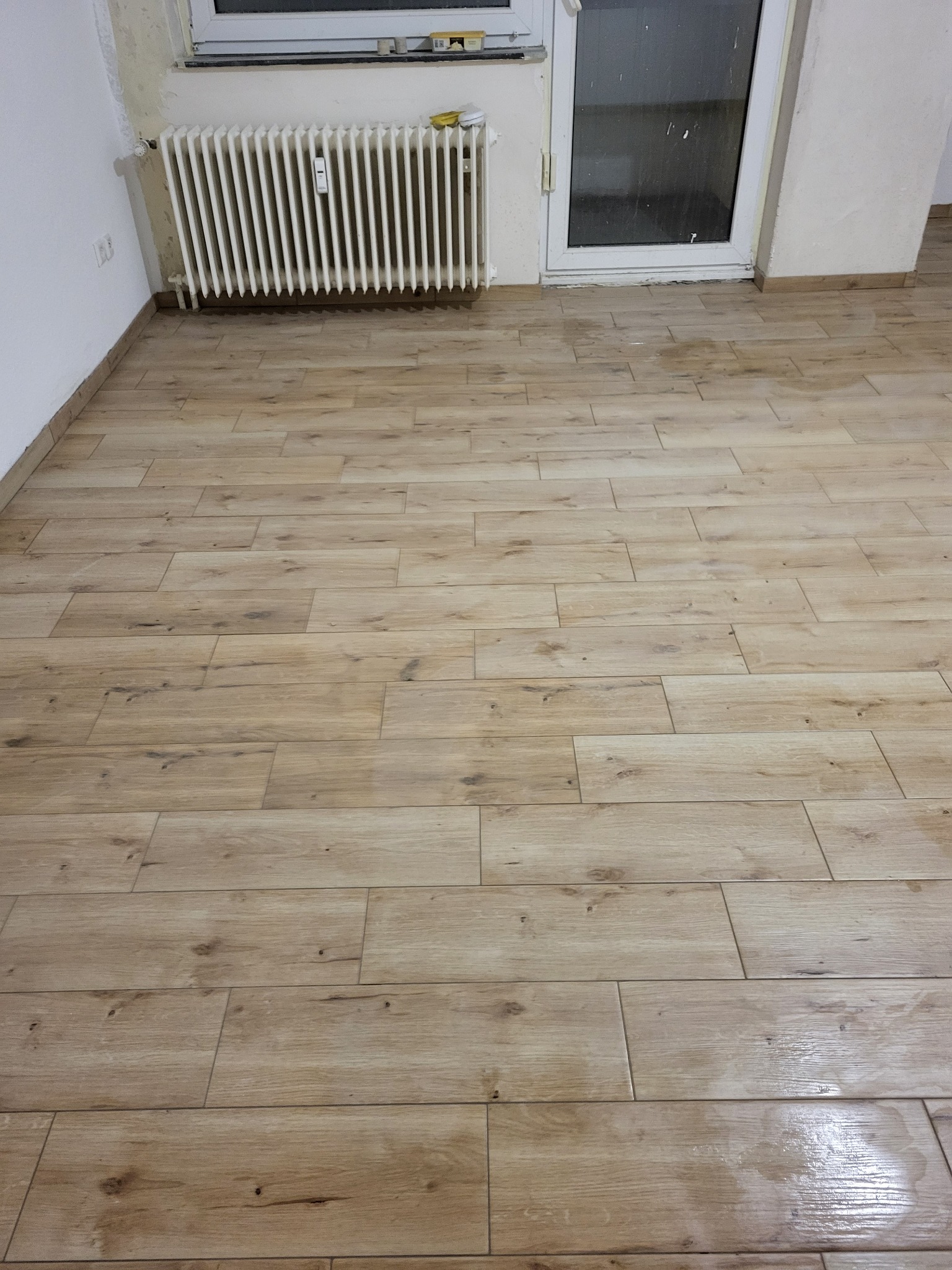 Ułożona podłoga z płytek imitujących drewno w pomieszczeniu z grzejnikiem i drzwiami balkonowymi, widoczne ślady po pracach wykończeniowych na ścianach.