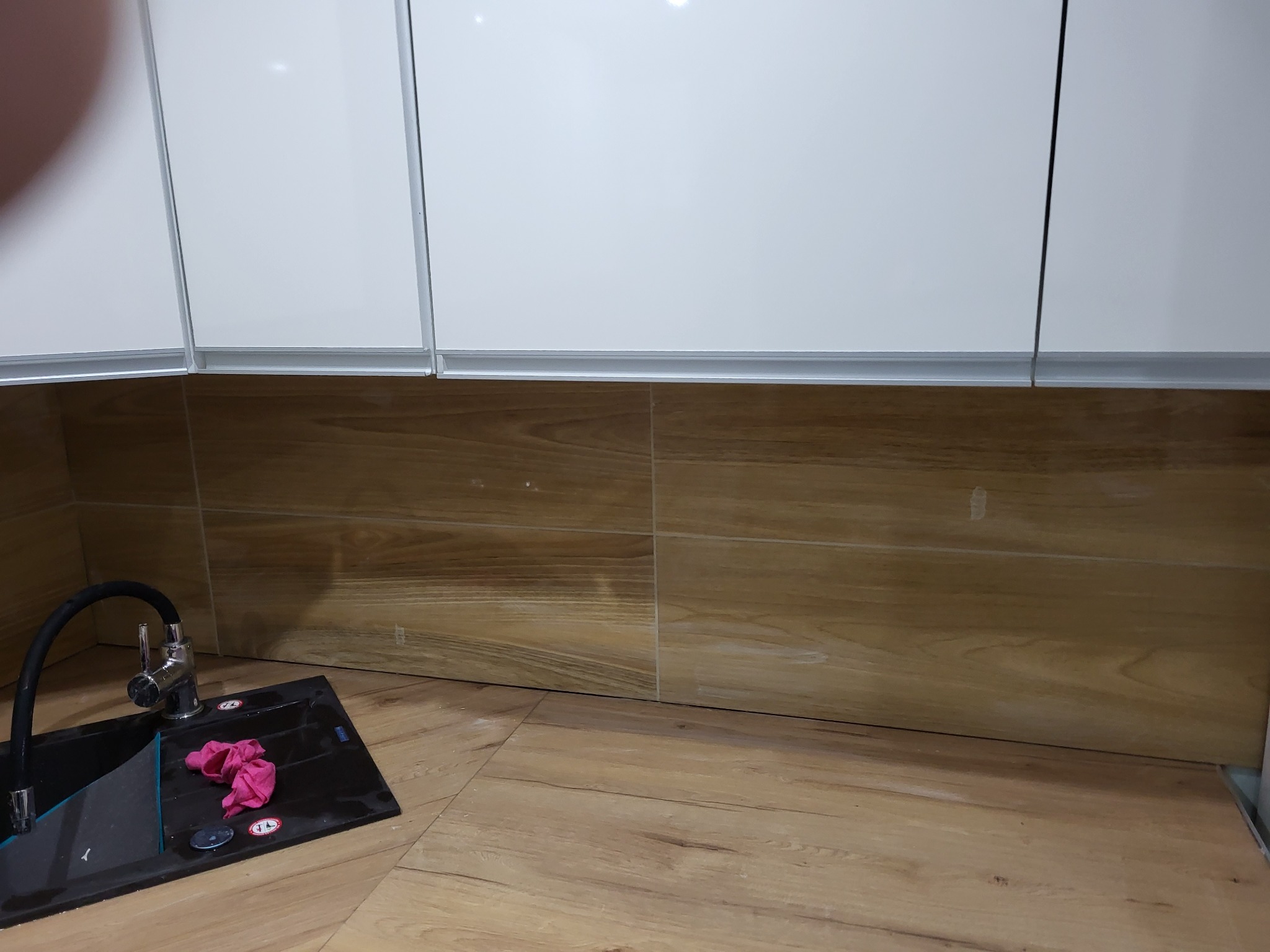 Nowoczesna kuchnia z białymi szafkami wiszącymi, drewnopodobnymi panelami ściennymi i podłogą, czarnym zlewem z chromowaną baterią i różową ścierką.