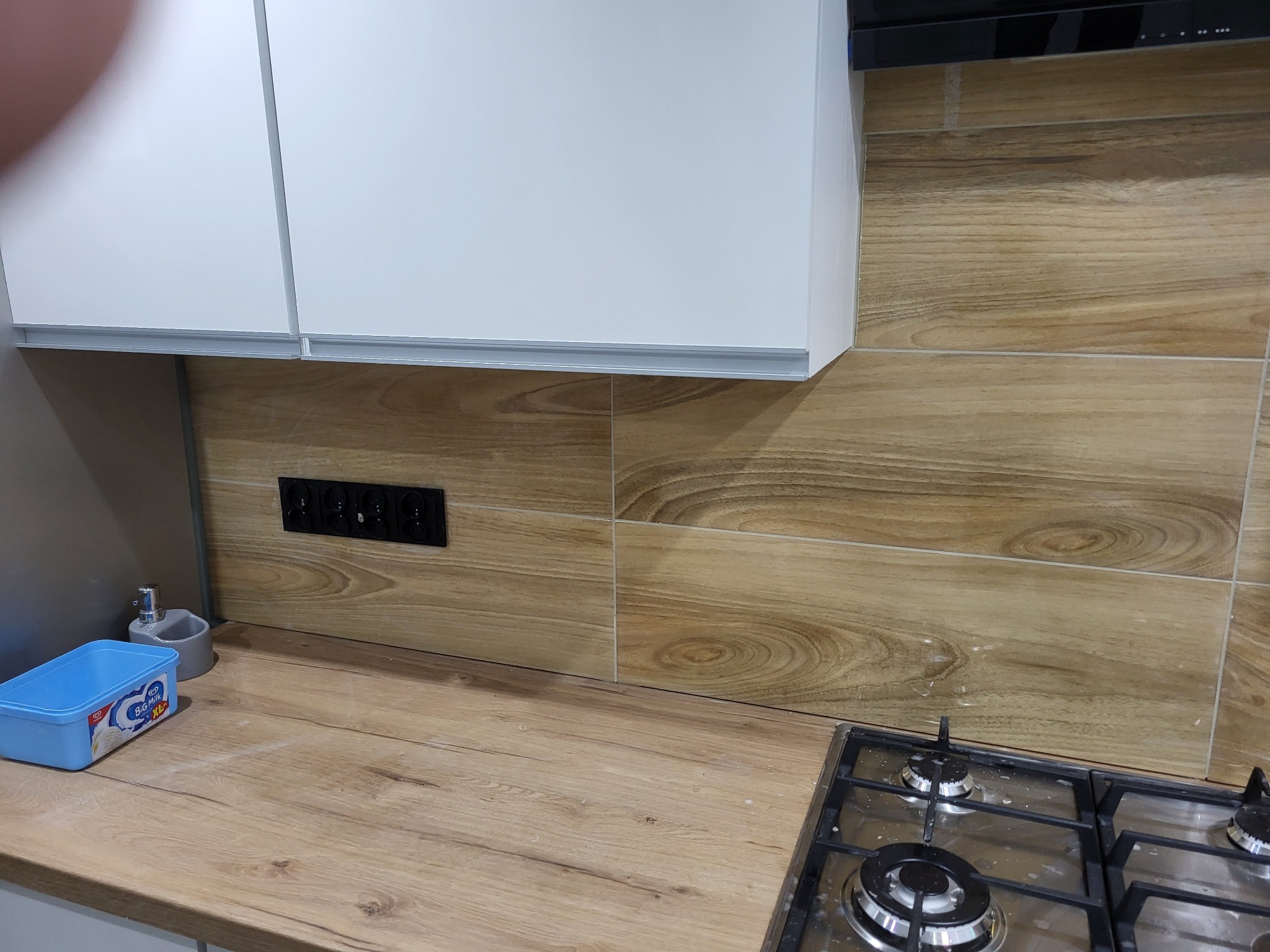 Fragment kuchni po remoncie: białe szafki wiszące, blat imitujący drewno, ceramiczne płytki ścienne z wzorem drewna, czarne gniazdka elektryczne, płyta gazowa, pojemnik na blacie.