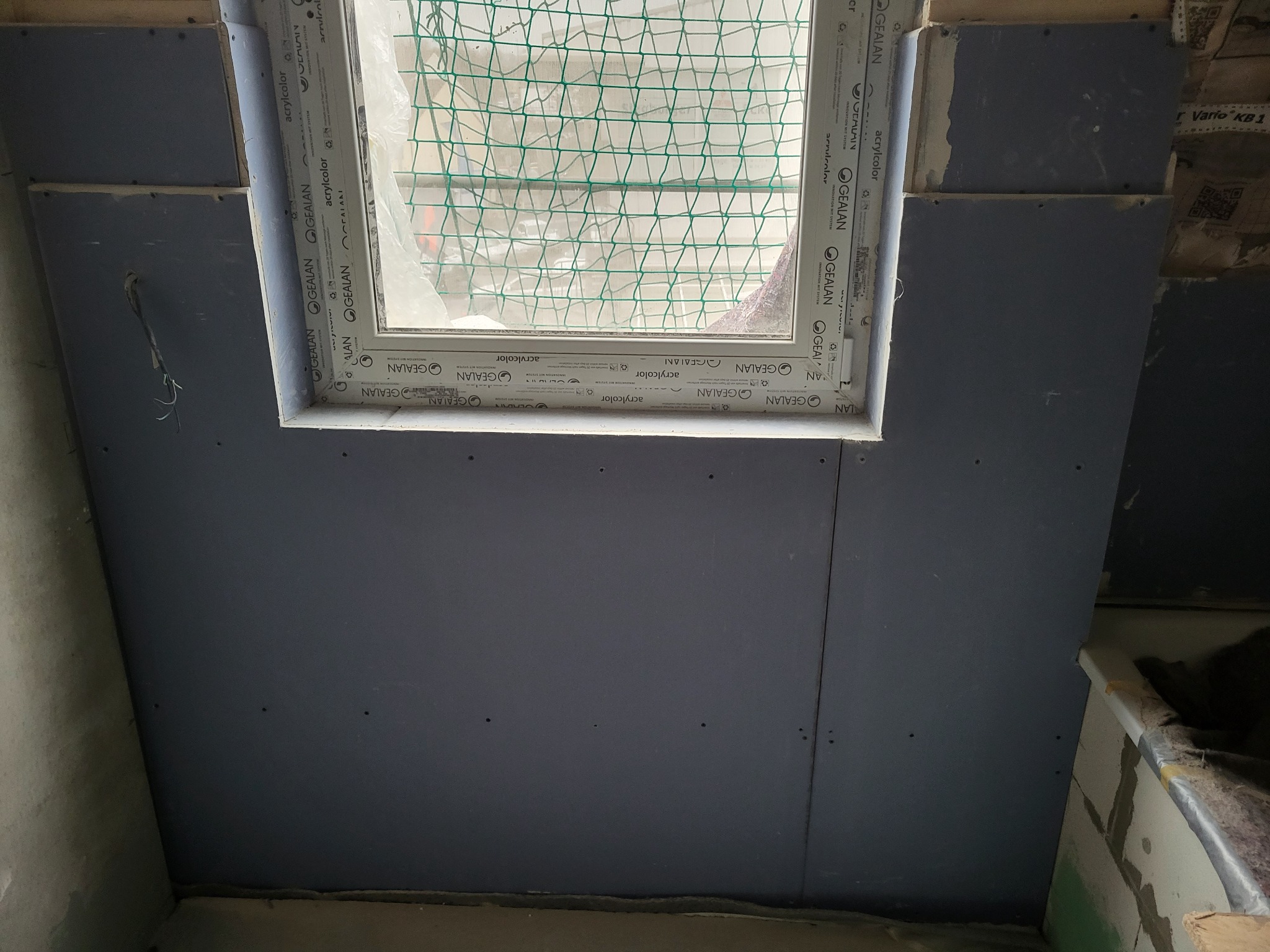 Montaż płyty gipsowo-kartonowej wokół okna dachowego, widoczna konstrukcja okna z szybą zabezpieczoną siatką ochronną, surowy stan wykończenia wnętrza.