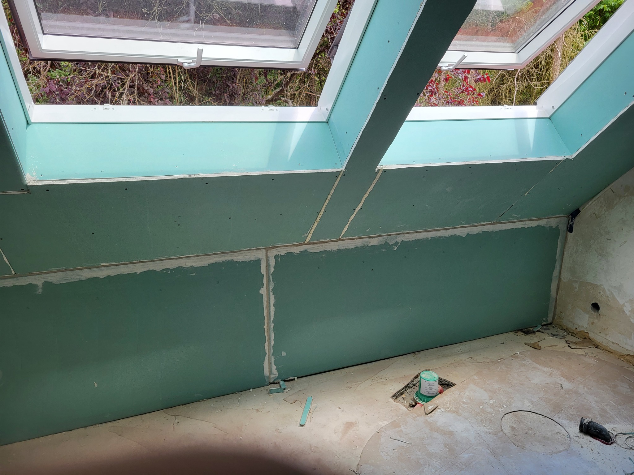 Płyty gipsowo-kartonowe na poddaszu z oknami dachowymi, widoczne łączenia płyt przed szpachlowaniem, fragment podłogi z wystającą instalacją hydrauliczną i resztkami materiałów budowlanych.