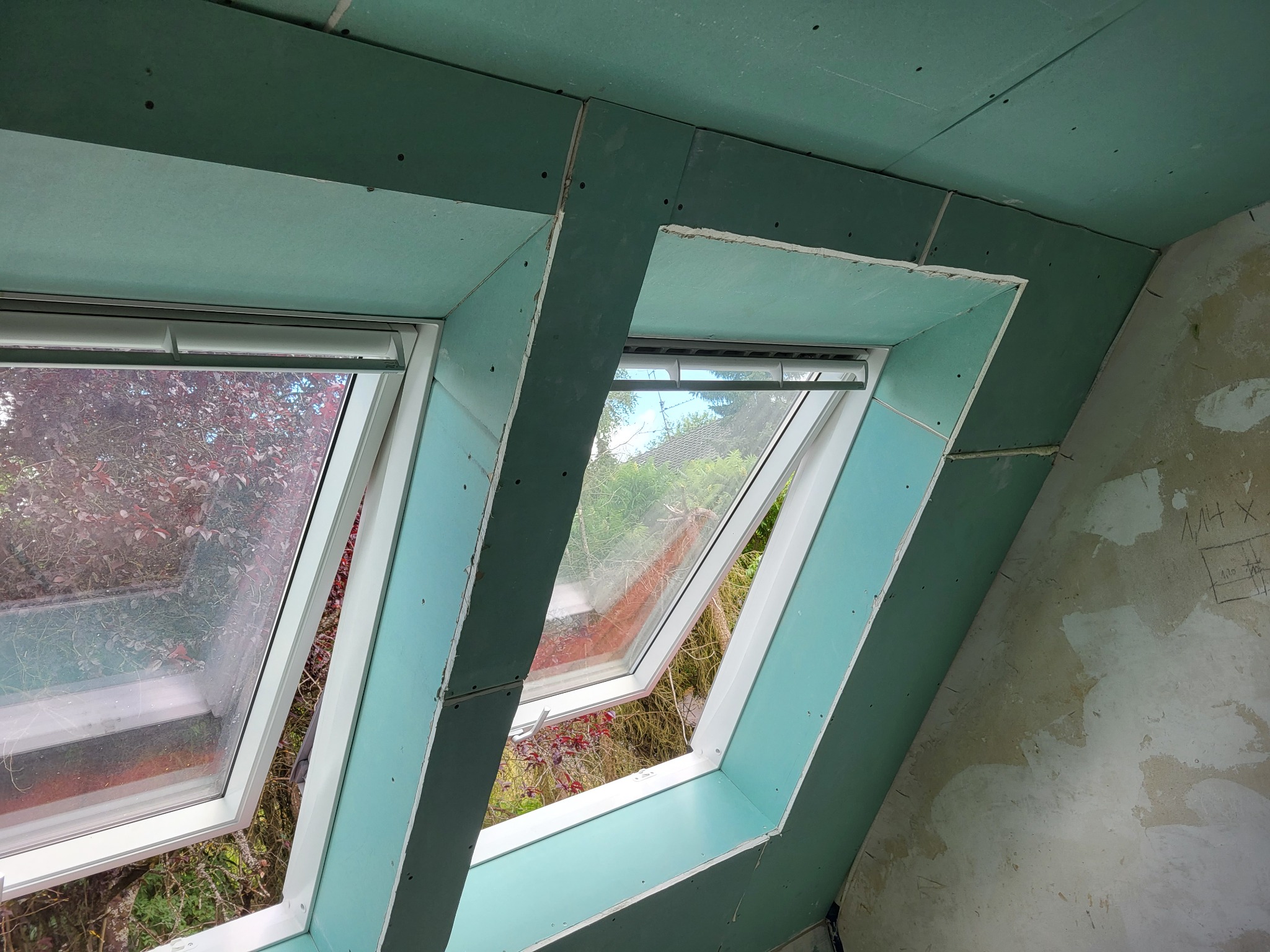 Dwa okna dachowe w trakcie montażu, widoczne płyty gipsowo-kartonowe i surowa ściana z odręcznymi notatkami.