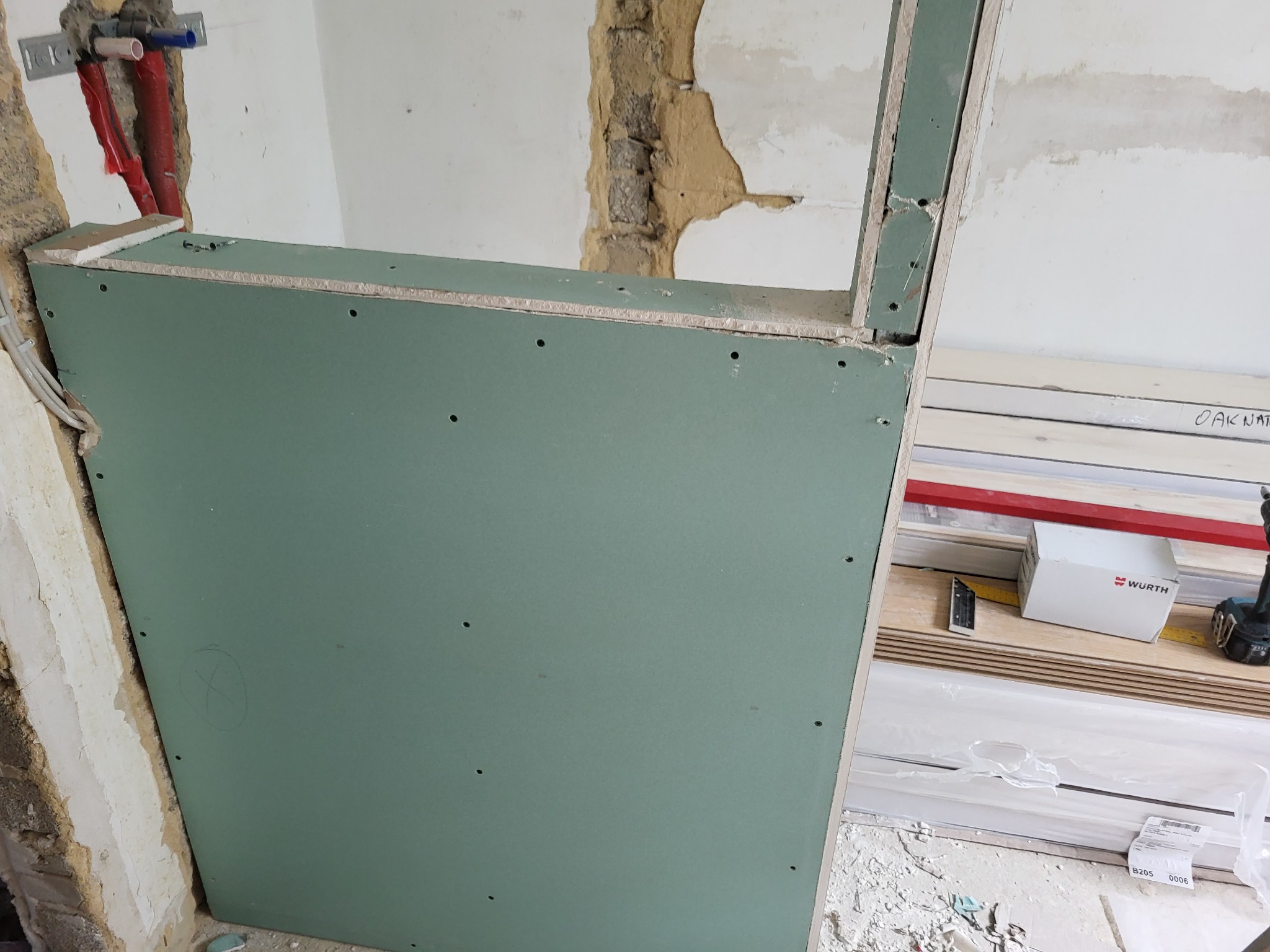 Fragment ściany z zamontowaną płytą gipsowo-kartonową w trakcie prac remontowych, widoczne instalacje hydrauliczne, surowa ściana i narzędzia pracy.