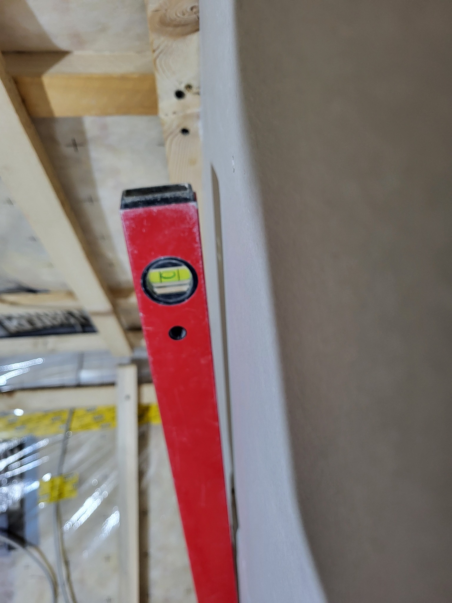 Czerwona poziomica pionowo przylegająca do świeżo nałożonej, jasnoszarej warstwy gładzi szpachlowej na ścianie, widoczna drewniana konstrukcja sufitu i folia ochronna w tle.